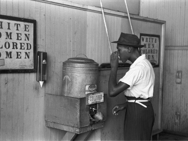 A photograph of a black man drinking from a water jug labeled "colored." This illustrates overt race discrimination.