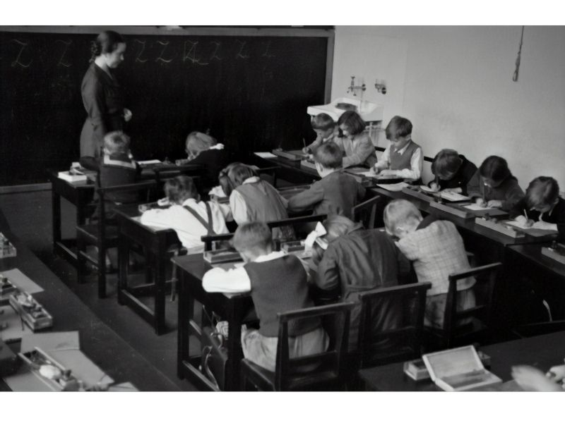Photo of 19th century classroom with students in rows of desks bent over doing their work